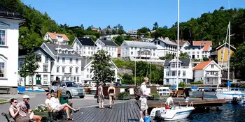 A serene harbor view of Tvedestrand with people sitting on a wooden dock near the water.